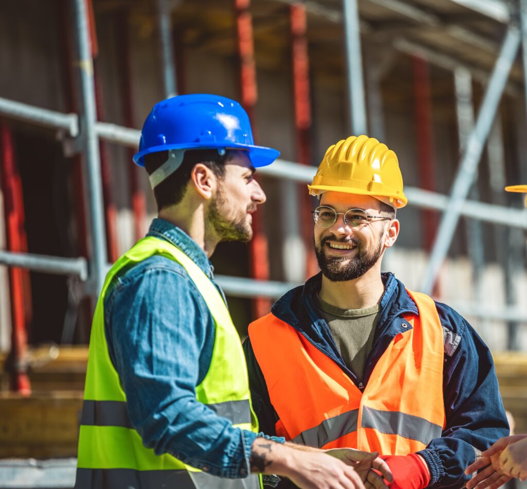 a group of three workers at a construction site wearing hard hats, safety glasses and reflective clothing, smiling and conversing.